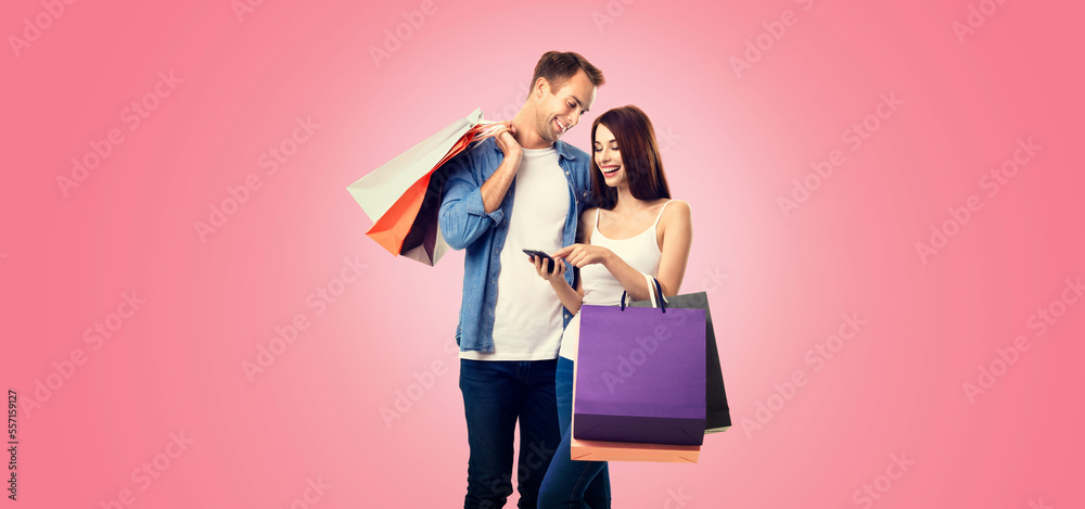 Holiday sales actions, rebates, discounts offers concept image - happy excited smiling couple with shopping bags, and cellphone, standing close to each other, on vivid rose pink background.