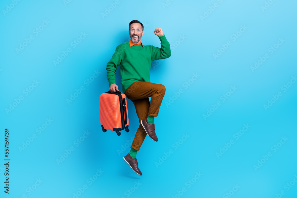 Full size photo of mature attractive male jumping raise fist hold baggage wear trendy green clothes isolated on blue color background Full size photo of mature attractive male jumping raise fist hold baggage wear trendy green clothes isolated on blue color background