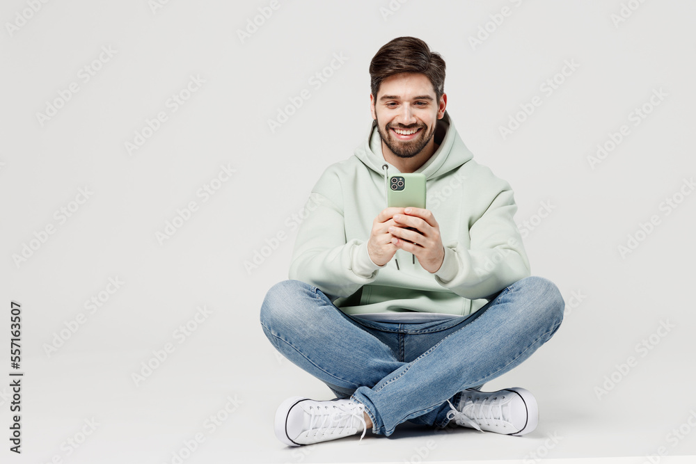 Full body smiling happy cheerful young caucasian man wear mint hoody look camera hold in hand use mobile cell phone isolated on plain solid white background studio portrait. People lifestyle concept. Full body smiling happy cheerful young caucasian man wear mint hoody look camera hold in hand use mobile cell phone isolated on plain solid white background studio portrait. People lifestyle concept.