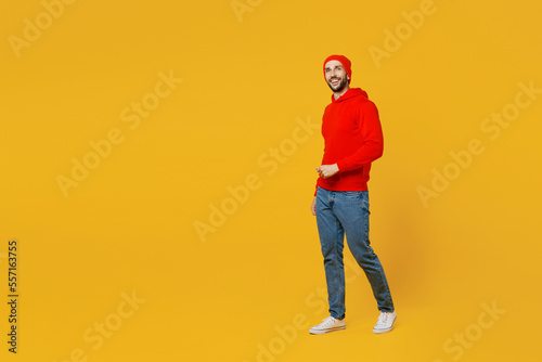 Full body side view smiling happy fun young caucasian man wears red hoody hat look camera walking going strolling isolated on plain yellow color background studio portrait. People lifestyle concept.