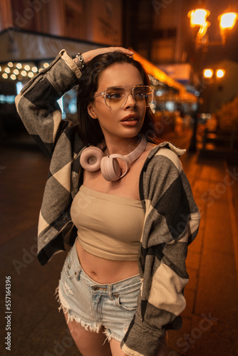 Beautiful woman with fashionable glasses and pink headphones in a fashion sweatshirt with a top and ripped jeans shorts walks in the night city with bokeh light