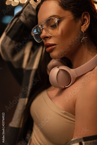 Street night woman portrait fashionable beautiful woman with stylish glasses and pink headphones in fashion trendy top and plaid shirt walking in the night city