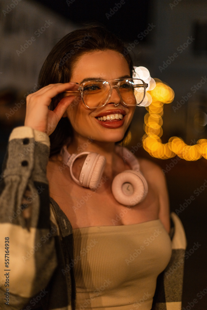 Happy beautiful fashionable woman with a smile with headphones wears stylish glasses in fashion casual clothes walks in the night city with bokeh lights