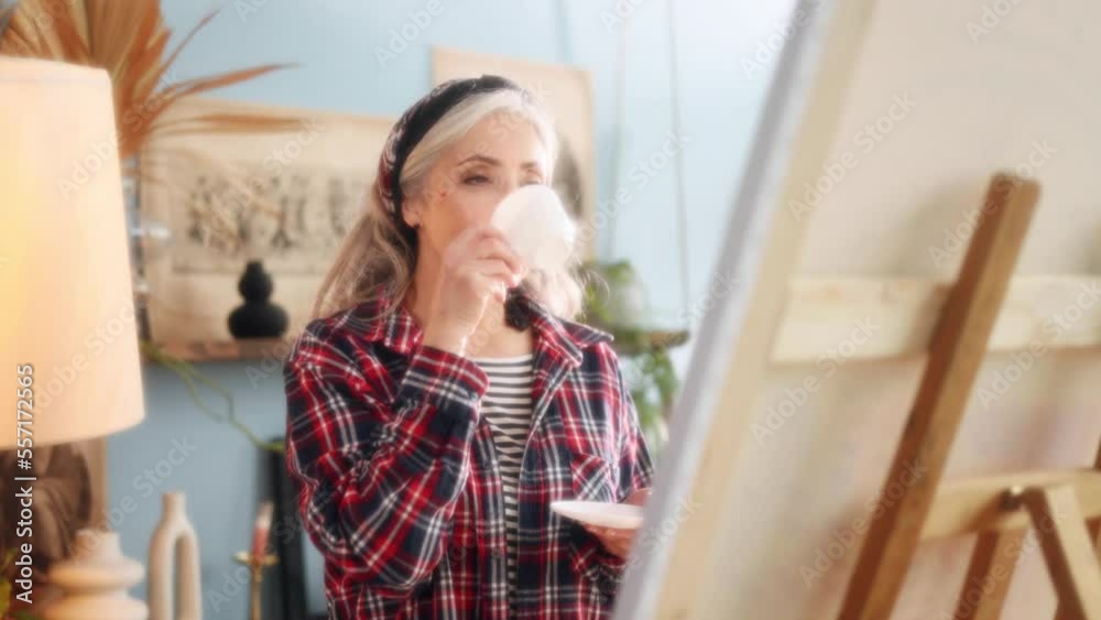 Dreamy soft portrait of charming stylish senior grey haired woman artist painter look at picture on canvas with cup of coffee or tea when finished artwork in the sunny creative workshop indoors Dreamy soft portrait of charming stylish senior grey haired woman artist painter look at picture on canvas with cup of coffee or tea when finished artwork in the sunny creative workshop indoors