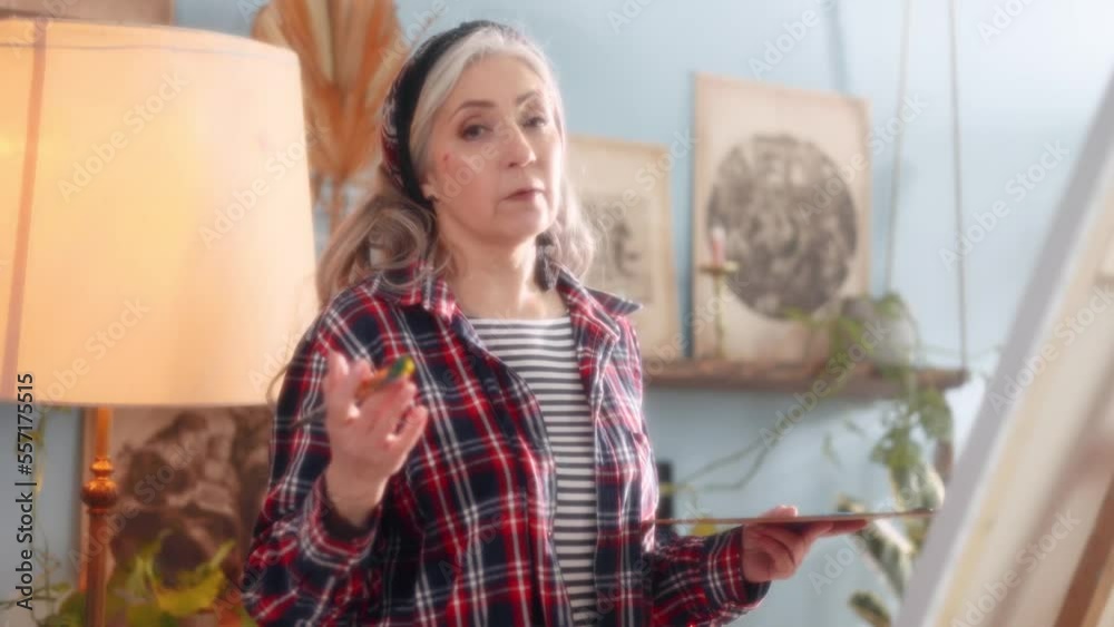 Dreamy soft shot of senior stylish grey haired woman teacher lecturer with brush in her hand talk at online class lesson in the creative workshop next to easel Distance art education Dreamy soft shot of senior stylish grey haired woman teacher lecturer with brush in her hand talk at online class lesson in the creative workshop next to easel Distance art education