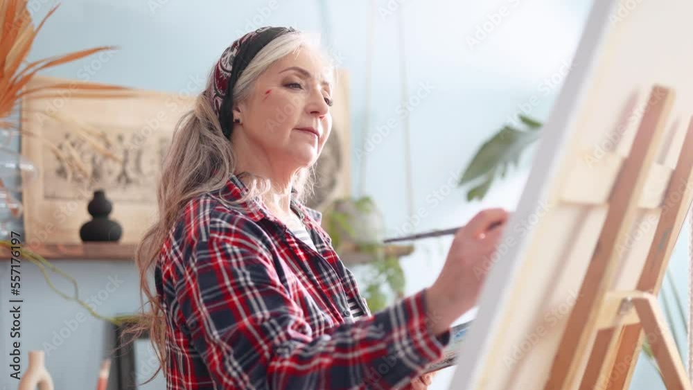 Portrait of charming senior woman artist painting picture on canvas creating artwork in the light sunny creative workshop Stylish mature grey haired lady enjoying leisure time with art hobby indoors Portrait of charming senior woman artist painting picture on canvas creating artwork in the light sunny creative workshop Stylish mature grey haired lady enjoying leisure time with art hobby indoors