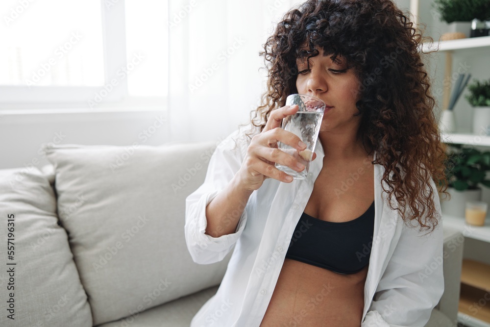 Pregnant woman smile and happiness taking pills and vitamins for pregnant women with pills hand water drinks clean water, maintaining water balance in the body, swelling, sitting on the couch at home Pregnant woman smile and happiness taking pills and vitamins for pregnant women with pills hand water drinks clean water, maintaining water balance in the body, swelling, sitting on the couch at home