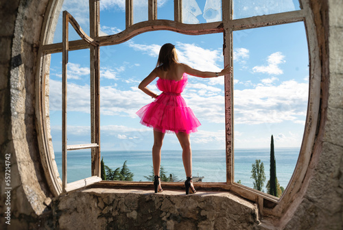 Sexy girl in a beautiful pink dress on the background of an old window in an abandoned castle