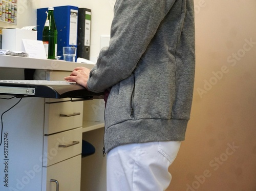 Schlanke junge Ärztin mit weißer Hose und grauer Jacke steht vor weißem Schubladenschrak und tippt auf Tastatur von Laptop in Arztpraxis