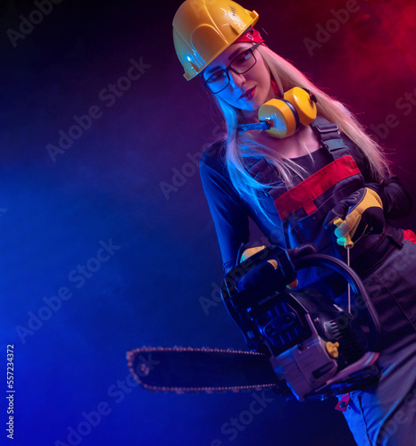 sexy girl with a chainsaw in a construction jumpsuit and helmet on a black background in neon light