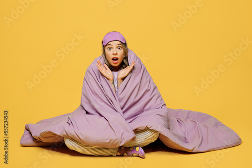 Full body young shocked confused woman she wears purple pyjamas jam sleep eye mask rest relax at home sit wrap covered blanket duvet spread hands isolated on plain yellow background Night nap concept © ViDi Studio