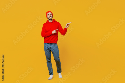 Full body fun young man wear red hoody hat point index finger aside indicate on workspace area copy space mock up isolated on plain yellow color background studio portrait. People lifestyle concept.