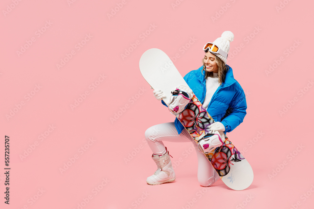 Full body snowboarder fun woman wear blue suit goggles mask hat ski padded  jacket kneeling hol snowboard isolated on plain pastel pink background  Winter extreme sport hobby weekend trip relax concept foto