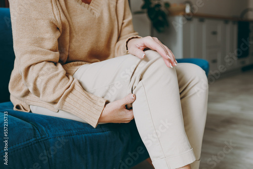 Cropped close up photo of pensioner woman wearing casual clothes sit on blue sofa hold knee suffer from leg knee pain inflammation stay home flat spend free spare time in living room indoor grey wall.