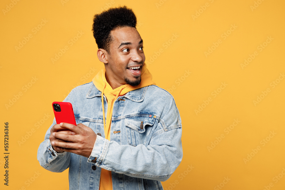 Young surprised shocked man of African American ethnicity wear denim jacket hoody hold in hand use mobile cell phone in red case look aside on area isolated on plain yellow background studio portrait.
