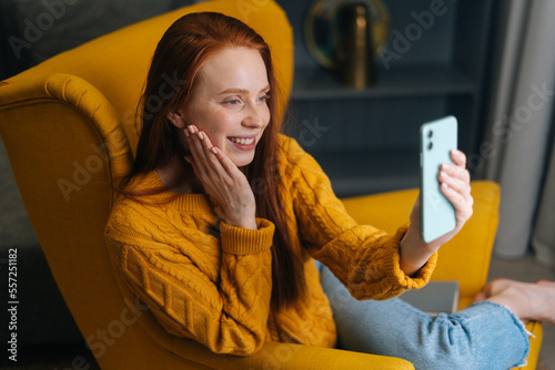 Pretty cheerful redhead young woman taking selfie picture, chatting by video call using smartphone sitting in cozy yellow armchair at home, smiling looking at camera. Lifestyle, social network concept