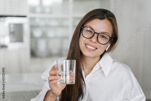 Beautiful Spanish young businesswoman in white shirt and glasses holds glass of water, toothy smiles looks at camera. Pretty brunette Italian woman offers pure water. Healthy lifestyle. Eco concept.
