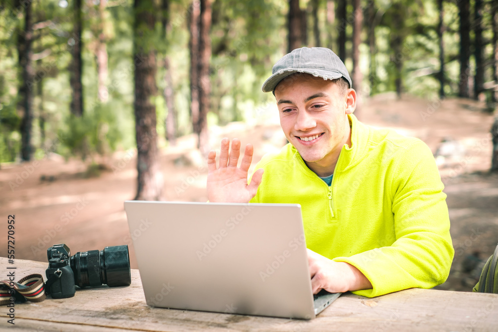 Young beautiful boy freelancer using video conference while working on laptop computer from remote. Happy traveler working distantly while enjoying nature mountain landscape during vacations