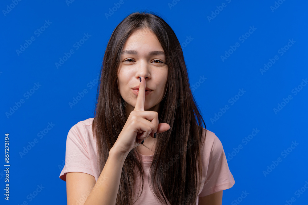Young woman holding finger on lips, blue studio background. Pretty teen girl with gesture of shhh, secret, silence, conspiracy, gossip concept Young woman holding finger on lips, blue studio background. Pretty teen girl with gesture of shhh, secret, silence, conspiracy, gossip concept