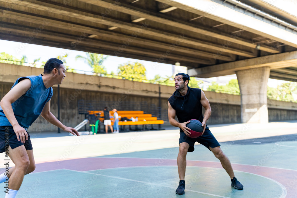 Two man athlete playing streetball match shooting and defense basketball on outdoors court together in sunny day. Sportsman do sport training basketball at street court under highway in the city.