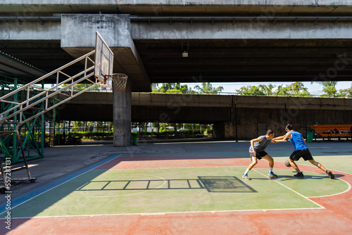 Two man athlete playing streetball match shooting and defense basketball on outdoors court together in sunny day. Sportsman do sport training basketball at street court under highway in the city. photo