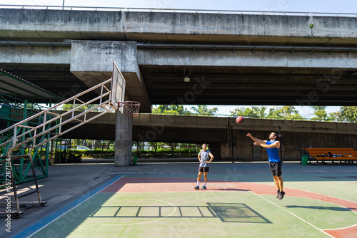 Two man athlete playing streetball match shooting and defense basketball on outdoors court together in sunny day. Sportsman do sport training basketball at street court under highway in the city.