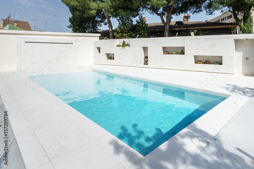A white marble pool filled with inviting chlorinated water, a garden with a lawn and trees and niches with flower pots on one wall