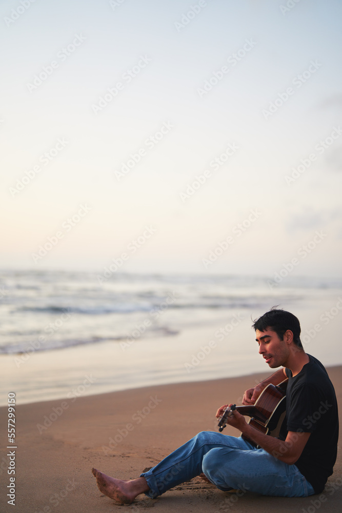 young latino male playing guitar and singing alone relaxed sitting on the beach shore at sunset