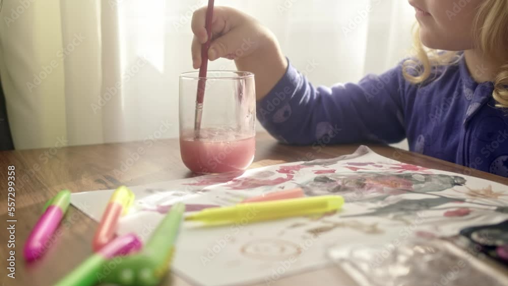 child rinses paint brush from red color in pink color water. Wash brush