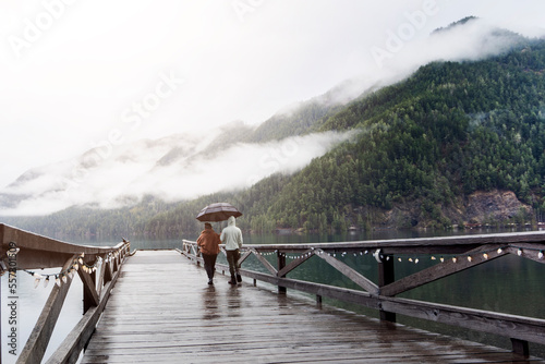 
A young sympathetic couple stands with an umbrella in their hands against the backdrop of a beautiful lake and mountains.