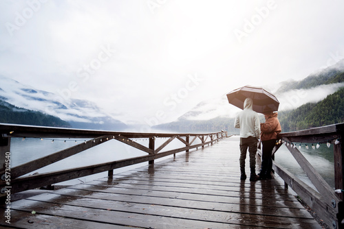 
A young sympathetic couple stands with an umbrella in their hands against the backdrop of a beautiful lake and mountains.