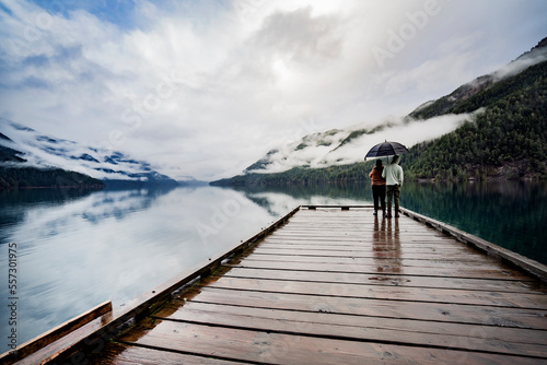 
A young sympathetic couple stands with an umbrella in their hands against the backdrop of a beautiful lake and mountains.