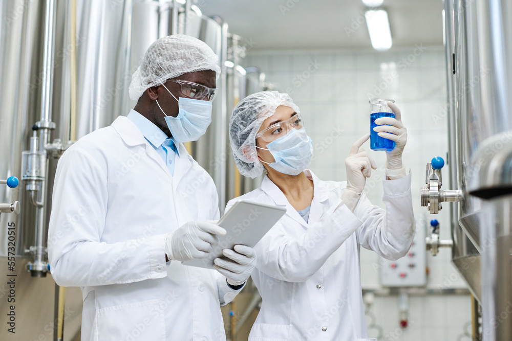 Multiethnic couple of scientists conducting research in team at factory, they examining blue liquid in flask