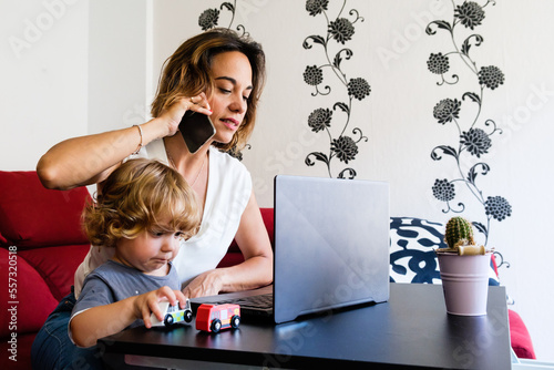 A single mother talks on the phone while working from home on her laptop and takes care of her young son, who plays with his toys. photo
