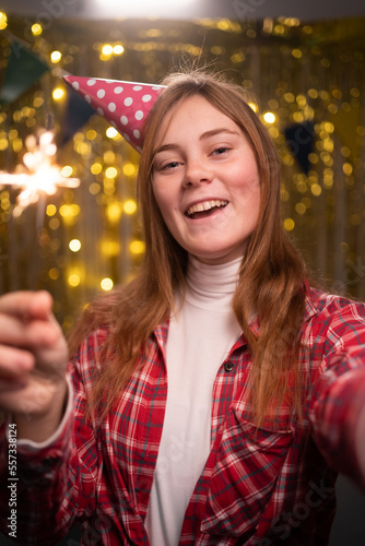 Happy positive blonde girl in party hat taking selfie with sparklers celebrating birthday at home