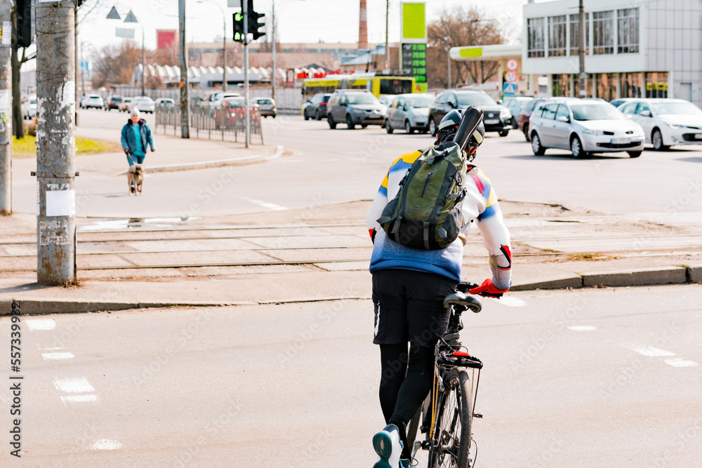Naklejka premium A man with a backpack and helmet crosses the road with a bike by pushing with one leg. Crosswalk. Pedestrian. Cross. Street. City. Outdoor
