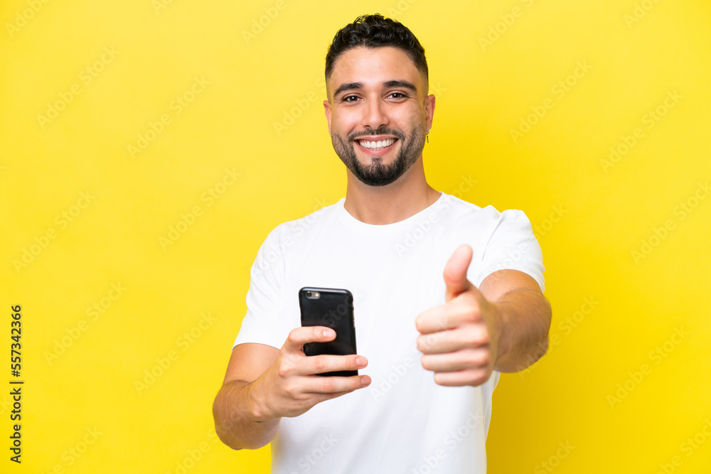 Young Arab handsome man isolated on yellow background using mobile phone while doing thumbs up Young Arab handsome man isolated on yellow background using mobile phone while doing thumbs up