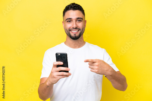 Young Arab handsome man isolated on yellow background using mobile phone and pointing it © luismolinero