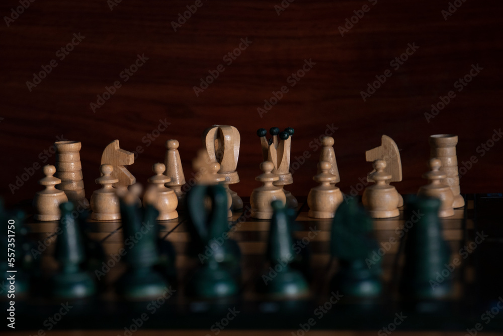 Chess board with white chess pieces in background and black chess pieces in foreground out of focus with wooden brown background. Concept: chess Chess board with white chess pieces in background and black chess pieces in foreground out of focus with wooden brown background. Concept: chess