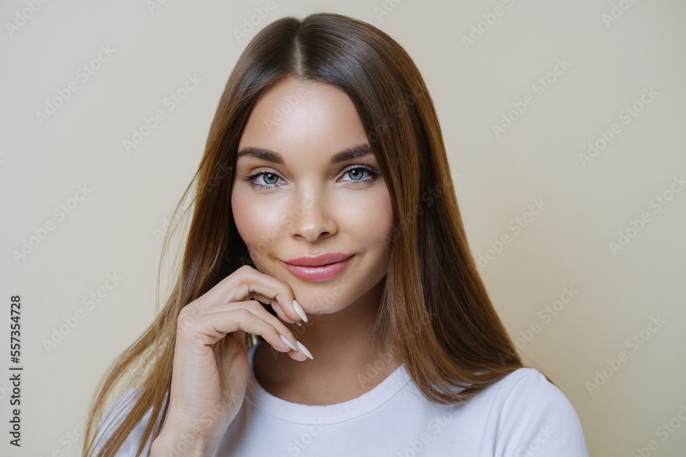 Charming good looking millennial woman keeps hand near face, has healthy glowing skin, dark hair, blue eyes, dressed in casual wear, isolated over brown background. Human face expressions concept Charming good looking millennial woman keeps hand near face, has healthy glowing skin, dark hair, blue eyes, dressed in casual wear, isolated over brown background. Human face expressions concept
