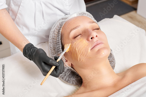 Cosmetologist applies honey mask on woman face for moisturize face skin, anti aging cosmetic procedure in beauty spa salon. Beautician in black gloves holds cosmetic brush for applying honey mask photo