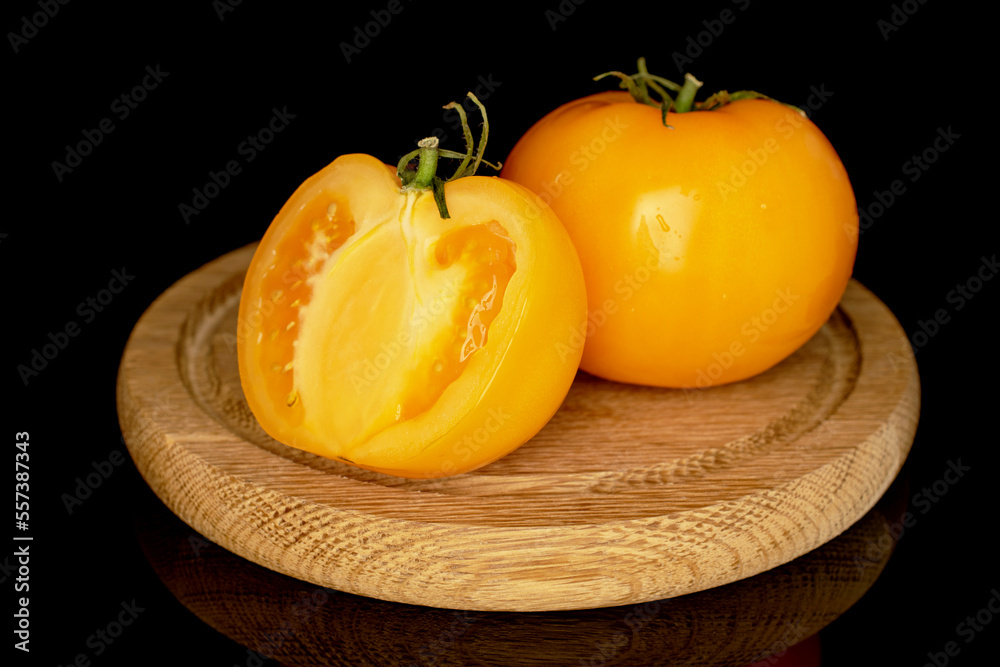 One whole and one half yellow organic juicy tomatoes on a wooden tray, close-up, isolated on black.