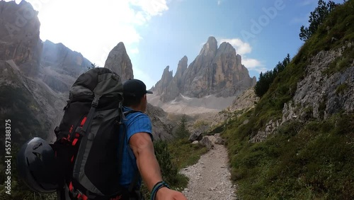 Man with big backpack taking a selfie while hiking along a narrow pathway in Italian Dolomites, South Tyrol (Alto Adige), Italy, Europe. Massive mountains with very steep sharp slopes. Raw landscape