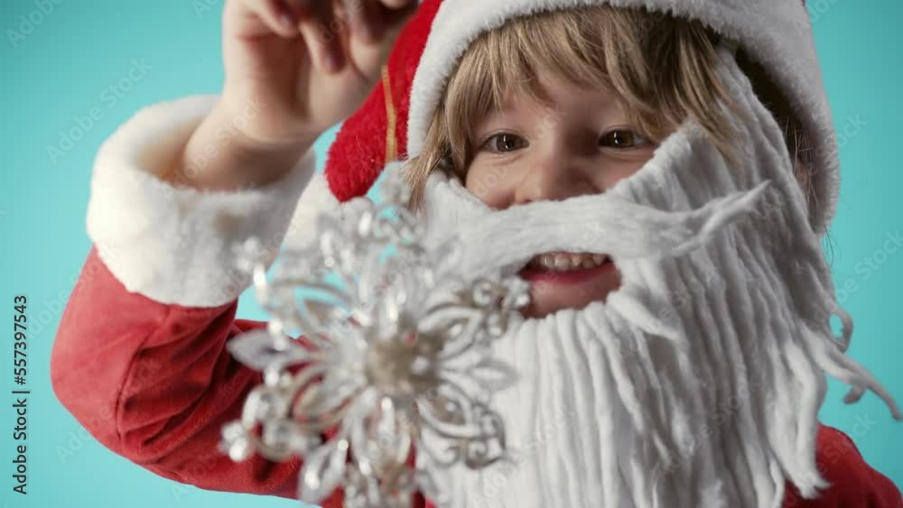 Lovely little child in Santa Claus costume, artificial beard playing with snowflake Christmas tree toy. Festive mood. Happy childhood, kids, funny son Lovely little child in Santa Claus costume, artificial beard playing with snowflake Christmas tree toy. Festive mood. Happy childhood, kids, funny son