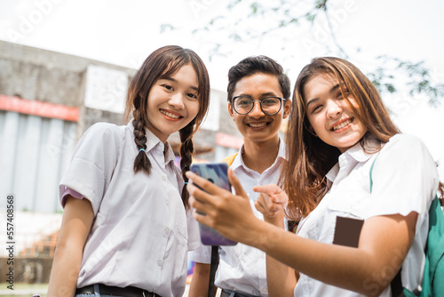 Boy student in glasses and two girls in uniform smiling while using smartphone