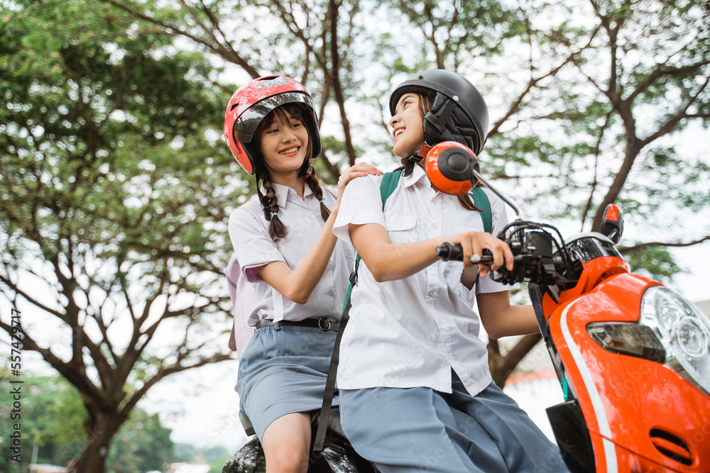 Two girls in high school uniforms wearing helmets chatting while riding a motorcycle together