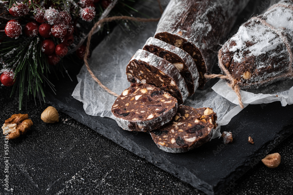 Chocolate salami filled with almonds, hazelnuts and raisins on the black background. Italian sweet sausage shaped dessert sprinkled with powdered sugar. Christmas and New Year festive pastry Chocolate salami filled with almonds, hazelnuts and raisins on the black background. Italian sweet sausage shaped dessert sprinkled with powdered sugar. Christmas and New Year festive pastry