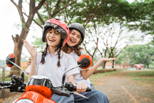 Two student girls wearing helmets smiling for the camera while riding motorbikes on the street