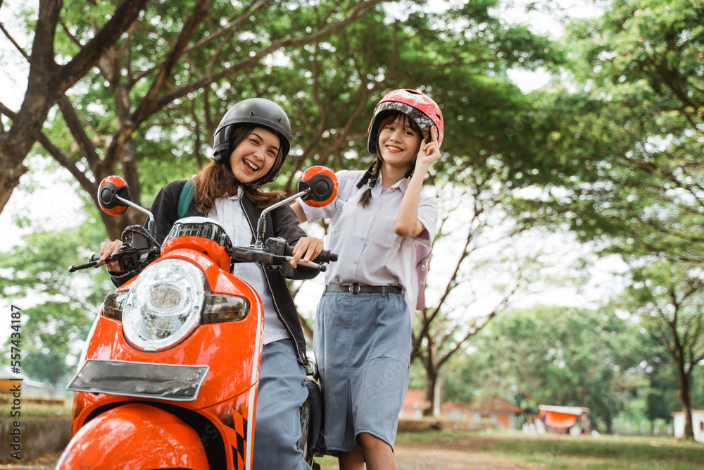 Two student girls wearing helmets and jackets smiling at the camera when they are going to ride a motorbike to school