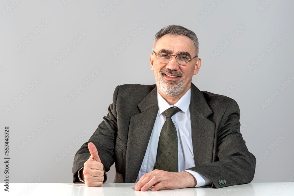 Adult man with thumb up wearing glasses in a formal suit with a tie. Smiling businessman with a beard sitting at the table in the office near the gray wall Adult man with thumb up wearing glasses in a formal suit with a tie. Smiling businessman with a beard sitting at the table in the office near the gray wall